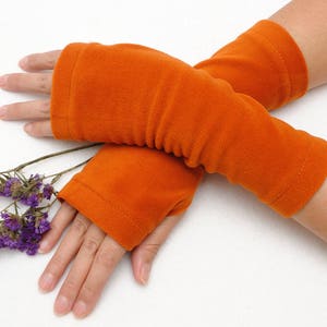 May include: A pair of orange fingerless gloves made of soft fleece fabric. The gloves are folded and placed on a white surface.