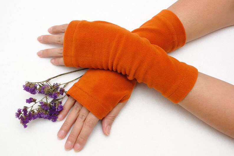 May include: A pair of orange fingerless gloves made of soft fleece fabric. The gloves are folded and placed on a white surface.