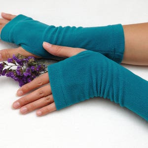 May include: A pair of teal blue fingerless gloves with a ribbed texture. The gloves are worn on a person's hands, and the person is holding a sprig of purple flowers.