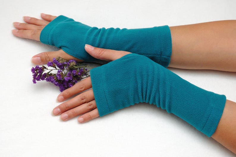 May include: A pair of teal blue fingerless gloves with a ribbed texture. The gloves are worn on a person's hands, and the person is holding a sprig of purple flowers.