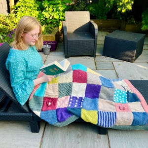 Könnte beinhalten: Eine Person sitzt auf einem Liegestuhl im Freien und liest ein Buch, während sie in eine bunte Patchwork-Strickdecke eingehüllt ist.