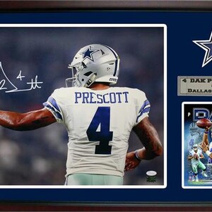 May include: A framed photo of a football player wearing a white jersey with the number 4 and the name "PRESCOTT" on the back. The player is wearing a blue helmet with a white star on it. The photo is signed by the player. The frame is navy blue with a silver plaque that reads "4 DAK PRESCOTT 4 QB DALLAS COWBOYS".