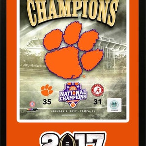 May include: Orange and black framed print of the 2016 National Champions Clemson Tigers football team. The print features the Clemson paw print logo, the team's score of 35, and the opponent's score of 31. The text "2016 National Champions" is also included. The print is framed in a black frame with a white mat.