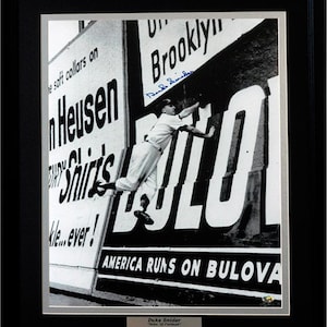 Puede incluir: Foto en blanco y negro de un jugador de béisbol, Duke Snider, saltando para alcanzar una valla publicitaria de relojes Bulova. La valla publicitaria dice "America Runs on Bulova".
