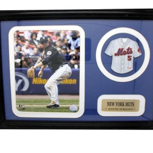 May include: A framed display featuring a baseball jersey and a photo of a baseball player in action. The jersey is white with blue stripes and has the word "Mets" and the number "5" on the front. The photo shows a player wearing a black jersey and white pants, holding a baseball glove and looking to the right. The frame is black and has a blue background with a gold plaque that reads "New York Mets - Player Signature".