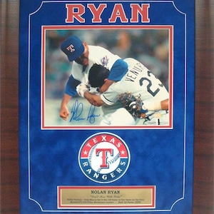 May include: A framed photo of Nolan Ryan, a baseball player, in a blue and white Texas Rangers uniform. The photo shows Ryan pitching and a player from the opposing team, wearing a white uniform with the number 23, is sliding into home plate. The photo is signed by Ryan and includes the Texas Rangers logo and the text "Nolan Ryan - "Earl"  The photo is framed in a brown wood frame.