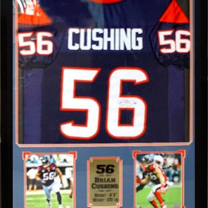 Op de afbeelding: Een ingelijst shirt met nummer 56 en de naam "CUSHING" op de rug. Het shirt is blauw met oranje accenten. Er zijn drie kleinere foto's van de speler die het shirt draagt tijdens een wedstrijd.