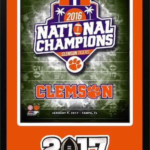 May include: A framed print of the 2016 Clemson Tigers National Champions. The print features a green and white football field with the team's logo and the words "National Champions" in purple and white. The print is framed in a black frame with an orange mat. The bottom of the print has the date "January 9, 2017" and the location "Tampa, FL".