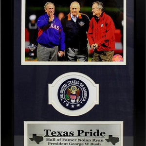 May include: A framed photo of three men, including Hall of Famer Nolan Ryan, President George W. Bush, and President George H.W. Bush, standing together at a baseball game. The frame features a silver plaque with the text "Texas Pride" and a small American flag patch.
