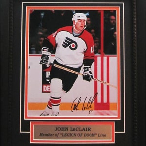 May include: A framed photo of John Leclair, a hockey player for the Philadelphia Flyers, wearing a white jersey with an orange and black logo. He is holding a hockey stick and is skating on the ice. The photo is signed by Leclair and includes the text "Member of "LEGION OF DOOM" Line".