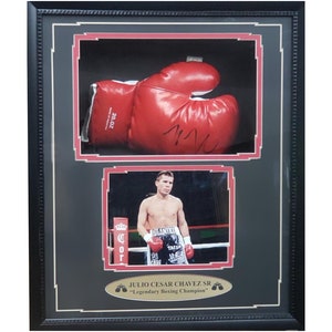 May include: A framed black and white photo of Julio Cesar Chavez Sr. in a boxing ring, with a red boxing glove signed "JCC" in the top frame. The text "JULIO CESAR CHAVEZ SR. "Legendary Boxing Champion" is below the photo.