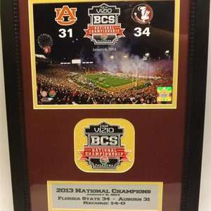 May include: A framed photo of the 2014 BCS National Championship game between Florida State and Auburn. The score is Florida State 34, Auburn 31. The photo is surrounded by a maroon mat and a black frame. The text "2013 National Champions" is printed on a silver plaque below the photo.