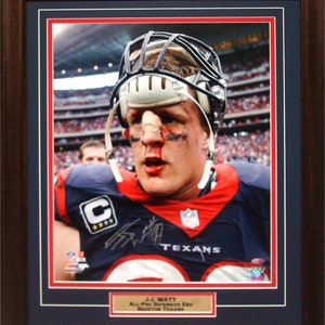 Op de afbeelding: Een ingelijste foto van J.J. Watt, een American football-speler voor de Houston Texans. Hij draagt een blauw en rood shirt met het teamlogo en een witte helm met een gezichtsmasker. Hij heeft een bloedneus en een snee in zijn gezicht. De foto is gesigneerd door J.J. Watt en bevat de tekst "J.J. Watt All-Pro Defensive End Houston Texans".