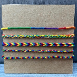 May include: A display of five colorful friendship bracelets. Each bracelet features a different design, including braided and chevron patterns, in a rainbow of colors. The bracelets are displayed on a brown cardboard backing.