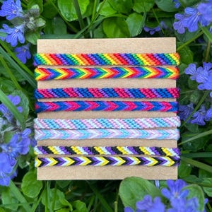 May include: Six colourful friendship bracelets with chevron patterns, presented on a brown card. The bracelets display rainbow, pink, blue, and purple hues. The background shows green grass and blue flowers, creating a natural setting.