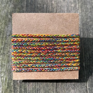 Könnte beinhalten: Ein Bündel aus farbenfrohem geflochtenem Kordel, das um eine rechteckige Kartonbasis gewickelt ist. Die Kordel weist einen Regenbogen aus Farben auf, darunter Rot, Gelb, Blau und Grün. Die Kordel ist eng gewebt und scheint aus einem weichen Material zu bestehen.