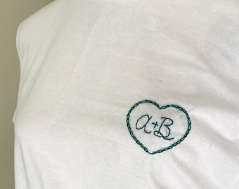 May include: White t-shirt with a teal embroidered heart design. The heart contains the letters "A+B" in a handwritten style. The shirt is made of a soft, lightweight fabric.