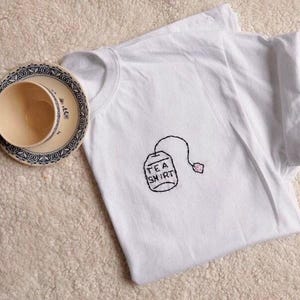 May include: White t-shirt with a hand-stitched tea bag design that reads "TEA SHIRT". A teacup and saucer with a blue and white floral pattern are next to the shirt. The teacup is beige.