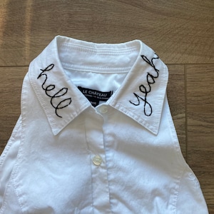 May include: White button-down shirt with a black embroidered collar. The collar has the words "hello" and "yeah" embroidered on it.