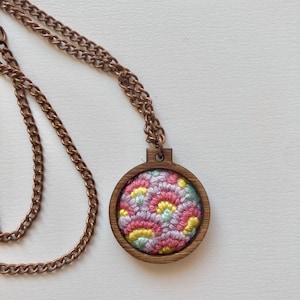 Puede incluir: Un collar con colgante circular que presenta un marco de madera oscura que contiene una pieza de tela con un bordado intrincado en tonos rosa, amarillo y azul claro. El colgante está unido a una cadena de metal color cobre con eslabones.