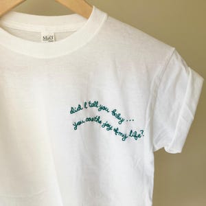 May include: A white short-sleeved t-shirt featuring dark green embroidered cursive text on the front right side that reads, Did I tell you baby... you are the joy of my life? The shirt has a crew neckline and is displayed on a wooden hanger.