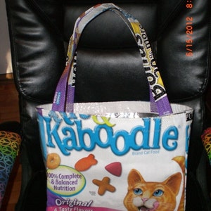 Op de afbeelding: Een witte tote bag met een blauw en geel ontwerp met de tekst "Kaboodle Brand Cat Food". De tas heeft een paarse en gele band en een afbeelding van een oranje kat met een roze neus en tong die uitsteekt. De tas heeft ook de tekst "100% Complete & Balanced Nutrition" en "Original 4 Tasty Flavors Chicken Liver".
