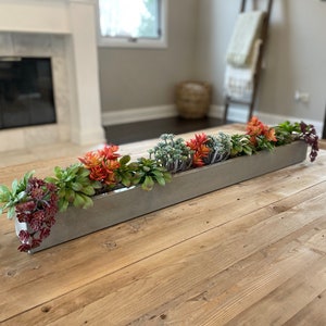 May include: A long, rectangular, gray concrete planter filled with a variety of succulents and small plants. The planter is sitting on a wooden table.