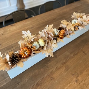 May include: A rectangular, light blue wooden centerpiece filled with fall-themed decorations. It features faux pumpkins in orange, gold, and white, along with pinecones, berries, and tan-colored leaves. The arrangement sits on a wooden table.