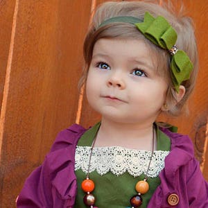 Könnte beinhalten: Ein junges Mädchen trägt ein grünes Kleid mit weißer Spitzenborte, einen lila Cardigan mit Knöpfen und eine bunte Perlenkette mit Quaste. Sie trägt auch ein grünes Stirnband mit Schleife.