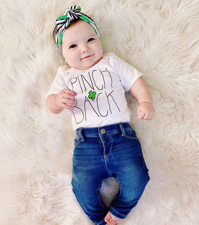 May include: A baby wearing a white onesie with the text "Pinch Back" and a green shamrock. The baby is also wearing a black and green headband and blue jeans.