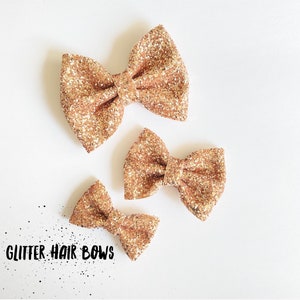 Puede incluir: Tres lazos para el pelo de purpurina dorada de diferentes tamaños. Los lazos están dispuestos en forma de triángulo sobre un fondo blanco. El texto "GLITTER HAIR BOWS" es visible en la esquina inferior izquierda de la imagen.