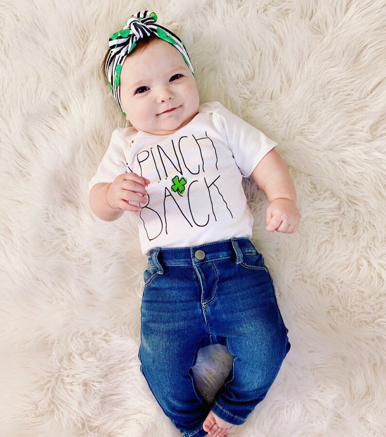 May include: A baby wearing a white onesie with the text "Pinch Back" and a green shamrock. The baby is also wearing a black and green headband and blue jeans.