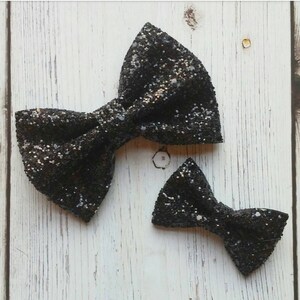 May include: Two black glitter bow ties, one larger than the other, on a white wooden background.