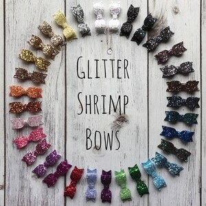May include: A collection of 20 glitter shrimp bows in various colors, including gold, silver, red, pink, blue, green, and purple. The bows are arranged in a circle on a white wooden background. The text "GLITTER SHRIMP BOWS" is written in black letters in the center of the circle.