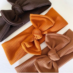 Pode incluir: Três faixas de cabelo de tecido em marrom, laranja e bege com um design de laço amarrado. As faixas são feitas de um tecido texturizado.