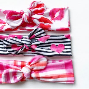 May include: Three pink and white patterned headbands with bows. The top headband has a red lip print, the middle headband has black and white stripes with pink hearts, and the bottom headband has a pink and white plaid pattern.
