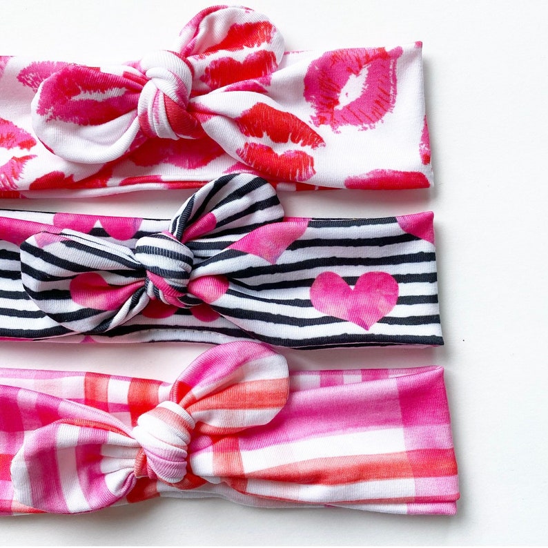 May include: Three pink and white patterned headbands with bows. The top headband has a red lip print, the middle headband has black and white stripes with pink hearts, and the bottom headband has a pink and white plaid pattern.