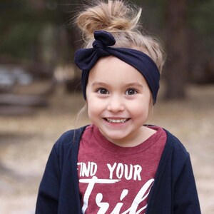May include: A young girl with a messy bun in her hair is wearing a navy blue headband with a bow. She is smiling and wearing a maroon t-shirt with the words "Find Your Tribe" printed on it. She is also wearing a navy blue cardigan.