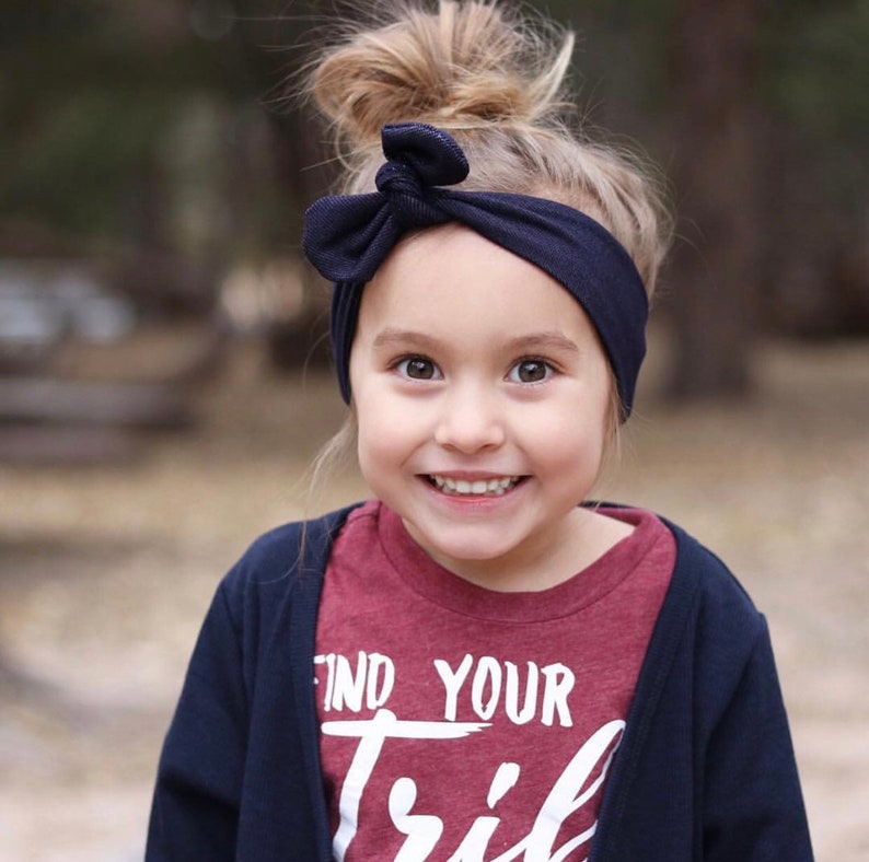May include: A young girl with a messy bun in her hair is wearing a navy blue headband with a bow. She is smiling and wearing a maroon t-shirt with the words "Find Your Tribe" printed on it. She is also wearing a navy blue cardigan.