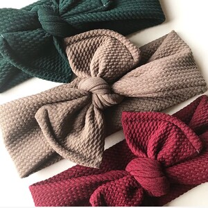 Pode incluir: Três faixas de cabelo de tecido em diferentes cores e estilos. A primeira faixa é verde escura com um laço. A segunda faixa é marrom com um laço grande. A terceira faixa é bordô com um laço menor.