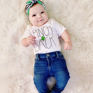May include: A baby wearing a white onesie with the text "Pinch Back" and a green shamrock. The baby is also wearing a black and green headband with a bow and blue jeans.