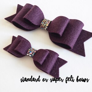 Könnte beinhalten: Zwei violette Filzbänder mit bunten Glitzerakzenten. Das kleinere Band liegt auf dem größeren Band. Der Text "standard or super felt bows" befindet sich am unteren Rand des Bildes.