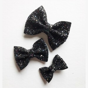 May include: Three black glitter bow hair clips of different sizes. The bows are made of sparkly black glitter fabric.