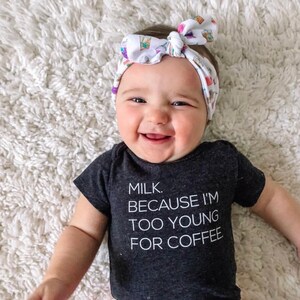 May include: A baby wearing a black bodysuit with the text "MILK. BECAUSE I'M TOO YOUNG FOR COFFEE." The baby is wearing a white headband with a colorful pattern. The baby is smiling and looking at the camera.