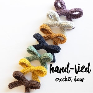 Puede incluir: Un conjunto de lazos de crochet hechos a mano en varios colores, incluyendo morado, gris, amarillo, verde, negro y marrón. Los lazos están dispuestos en una línea, con el lazo más grande en la parte superior y el lazo más pequeño en la parte inferior. El texto "lazo de crochet hecho a mano" está escrito debajo de los lazos.