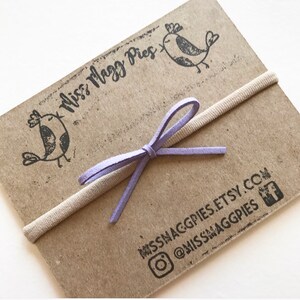 May include: A purple suede bow tied around a beige elastic headband. The headband is on a brown card with the text "Miss Magg Pie's" and the shop's social media handles.
