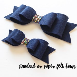 Peut inclure: Deux nœuds en feutre bleu marine avec des accents de paillettes colorées. Les nœuds sont noués au centre et ont des extrémités pointues. Le texte "standard or super felt bows" est écrit sous les nœuds.