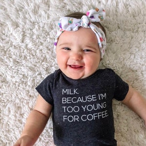 May include: A baby wearing a black bodysuit with the text "MILK. BECAUSE I'M TOO YOUNG FOR COFFEE." The baby is wearing a white headband with a colorful pattern. The baby is smiling and looking at the camera.