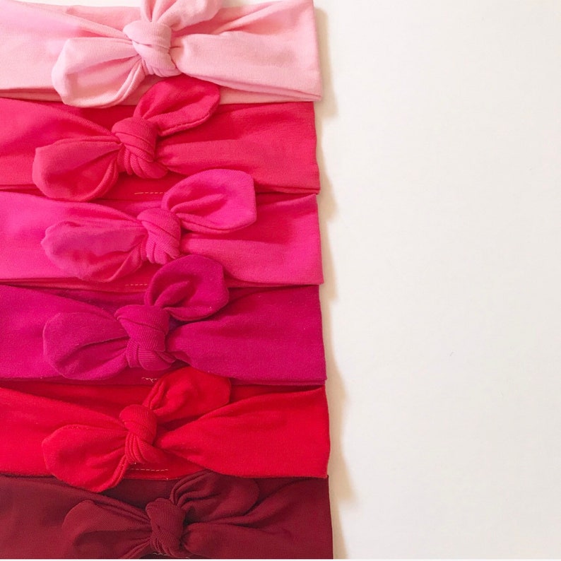 May include: Six headbands in various shades of pink and red, each with a knotted bow in the center. The headbands are arranged vertically, with the lightest shade at the top and the darkest shade at the bottom.