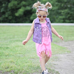 May include: A young girl wearing a pink dress with a denim vest and a floral headband. The dress has a fringe hem and the vest has distressed details. The girl is wearing sunglasses and has her hair in pigtails. The text on the dress says "You're really pretty".